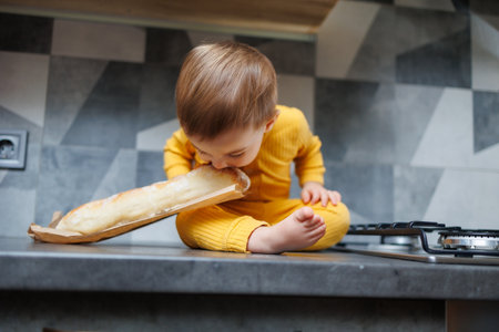 A cute 1-year-old boy in yellow clothes is sitting in the kitchen on the table with a fresh wheat baguette. Portrait of a cute one-year-old boy eating delicious breadの写真素材