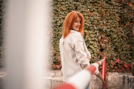 A pretty redhead young woman is walking down the street, she is dressed in jeans and a beige shirt....