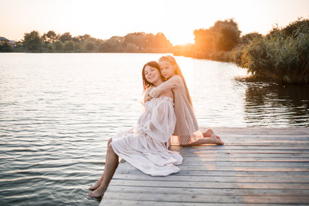 A pregnant woman with her daughter against the background of water at sunset, a woman with a belly enjoys the sunset by the riverの写真素材