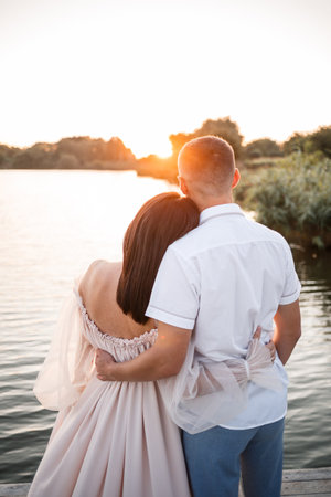 A pregnant woman and her husband stand on the pier and look at each other, a beautiful river and sunset, future parents spend time outdoors, a beautiful young motherの写真素材