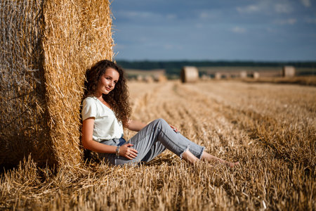 Beautiful young woman near a sheaf of hay in a field. Holidays in the village, a girl enjoying...