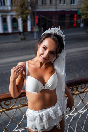 A beautiful young woman in exquisite lingerie stands on the balcony of a high-rise building. Morning of the bride on the wedding day.の写真素材