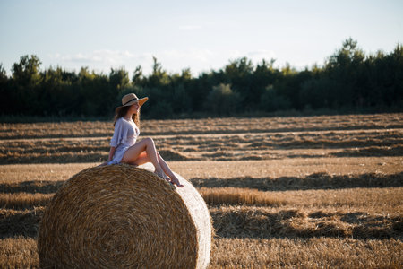 A beautiful young woman in a hat and a summer dress sits on a sheaf of hay in a field. Rural nature, wheat fieldの写真素材
