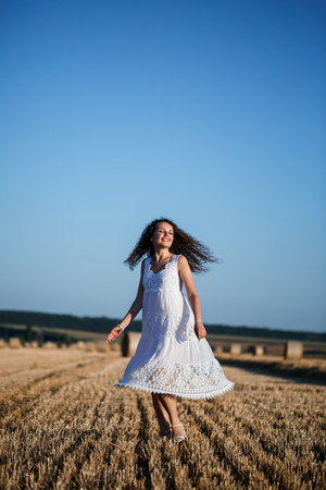 A young beautiful woman in a white summer dress stands on a mown wheat field with huge sheaves of hay, enjoying nature. Nature in the village. Selective focusの写真素材