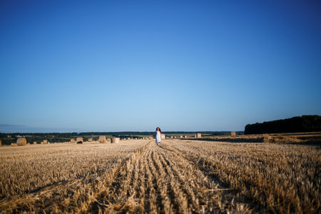 A young beautiful woman in a white summer dress stands on a mown wheat field with huge sheaves of...