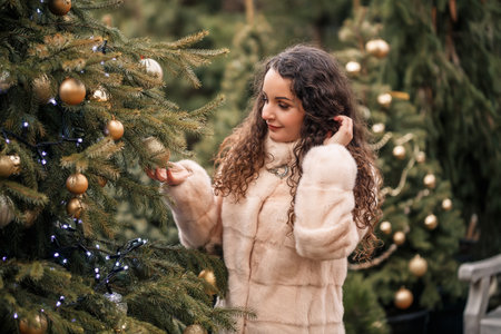 Smiling happy curly-haired woman in a fur coat strolls through the Christmas tree fair. Waiting for Christmas and New Year.の写真素材