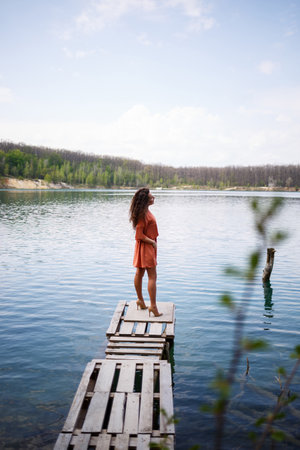 Wooden masonry. A girl with long wavy curly hair in an orange guipure dress and shoes on nature, in a forest against a lake, stood near trees and bushes. Young woman smiles and enjoys lifeの写真素材