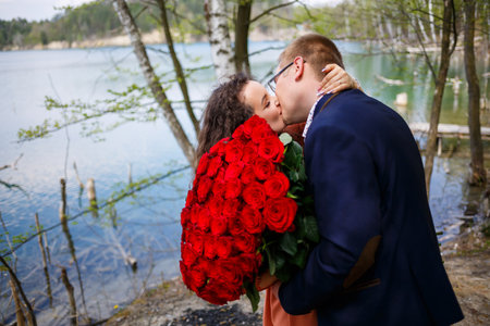 Romantic meeting of young people. A young woman agreed to marry her man. A guy in a suit with a bouquet of red roses gives the girl a bouquet, and they kiss in the forestの写真素材