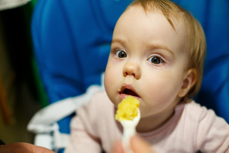 Photo portrait of a baby girl with pink cheeks she sits in a chair for feeding and eats porridge. Child sitting at homeの写真素材