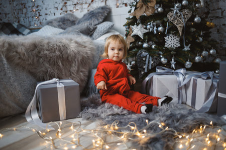 A little girl in a red warm sweater sits under a Christmas tree with toys and gifts. Happy childhood. New Year holiday atmosphereの写真素材