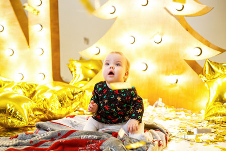 Little girl sitting on the floor against the background of a yellow Christmas tree. Happy child. New...