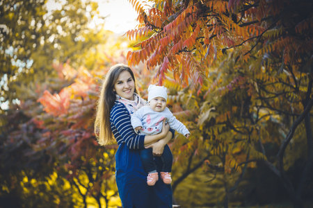 Beautiful woman in a dress plays with a child in her arms, standing in autumn nature with fallen...