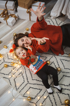 Mom with daughter in red warm sweaters are jumping on the bed. Happy motherhood. Warm family relationships. Christmas and New Year's interior. Love. Family concept.の写真素材