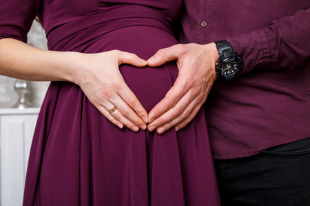Cropped image of husband holding belly of his pregnant wife making symbol heart hands. Pregnant...