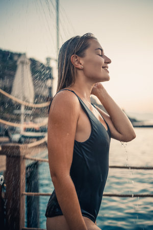 A beautiful young attractive woman in a black swimsuit with a beautiful figure stands under a summer shower on the pier by the sea. She is enjoying her vacation. Selective focusの写真素材