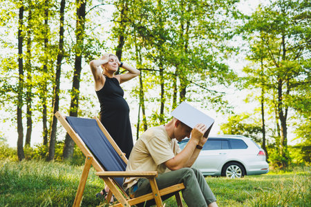 A family couple, a man and a woman, are relaxing in nature and working remotely with a laptop. Picnic of loving couples with work online. A pregnant woman rests in nature with her husbandの写真素材