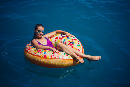 A young beautiful girl in a bright swimsuit lies on a large inflatable ring and floats on the blue...