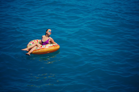 A young beautiful girl in a bright swimsuit lies on a large inflatable ring and floats on the blue sea on a bright sunny summer dayの写真素材