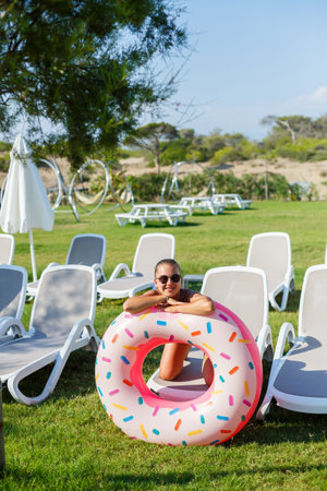 A young woman in a stylish swimsuit and sunglasses holds an inflatable donut ring in her hand. Beautiful happy girl posing and having fun in the sun. Vacation. travelの写真素材