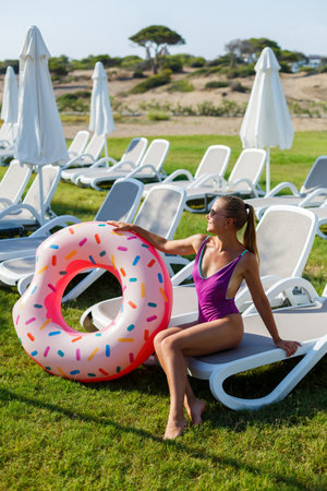 A slender young woman holds an inflatable circle in her hands, standing on the green grass. Attractive model with perfect body. Vacation concept, girl goes swimming in the seaの写真素材