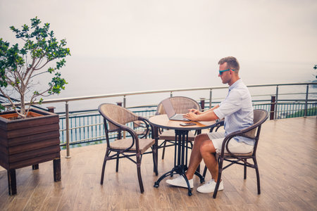 Handsome successful young male businessman sitting at a table by the pool with a laptop overlooking the Mediterranean Sea. Remote work on vacation. Vacation conceptの写真素材