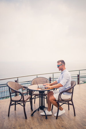 Handsome successful young male businessman sitting at a table by the pool with a laptop overlooking the Mediterranean Sea. Remote work on vacation. Vacation conceptの写真素材