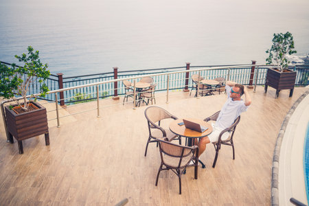 Handsome successful young male businessman sitting at a table by the pool with a laptop overlooking the Mediterranean Sea. Remote work on vacation. Vacation conceptの写真素材