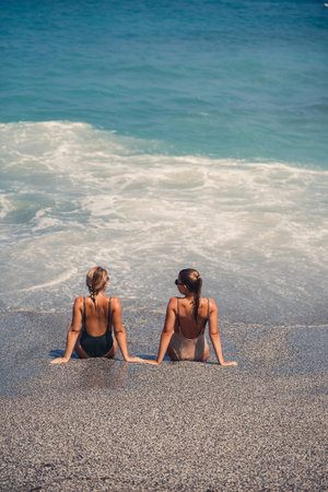 Two girls girlfriend are sitting on the sea sandy shore and the waves soaked them in bathing suits on a sunny warm dayの写真素材