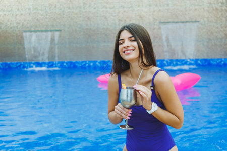 Cute woman in a swimsuit sitting by the pool and drinking a delicious cocktail and sunbathing by the...