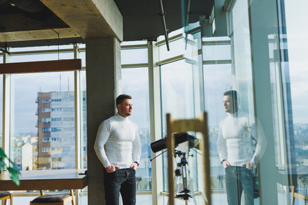 A young handsome man in a white sweater is standing in a spacious bright office. The concept of a...