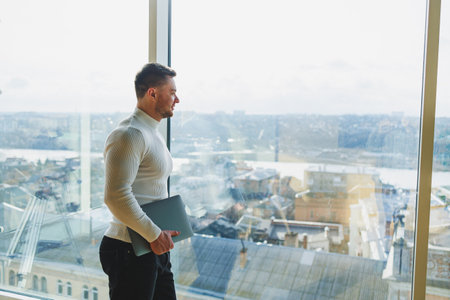 A young man in a modern workspace stands near a large window. A man in casual clothes with a laptop...
