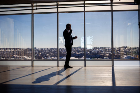 Rear view of a businessman looking out of a large window overlooking the city. He has a phone in his...