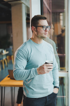 Young promising male manager in casual clothes standing by the window in the workspace and looking...