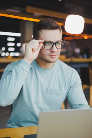 A young promising man in casual clothes sits at a table and works on a laptop. Work space for remote...