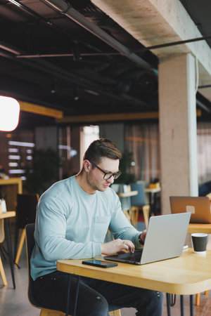A young promising man in casual clothes sits at a table and works on a laptop. Work space for remote...