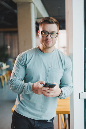 Young promising male manager in casual clothes standing by the window in the workspace and looking...