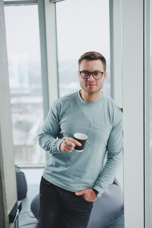 Smiling man in casual clothes standing near window with coffee in modern work area with big window during daytime.の写真素材