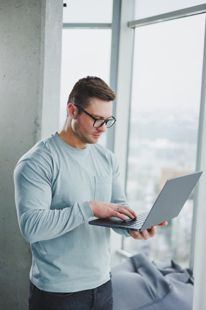Modern male manager in casual clothes stand by the window in the workspace with a laptop. Male...