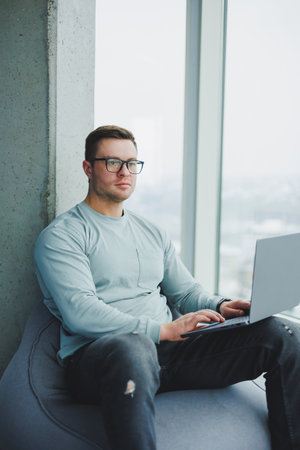 A young manager man in glasses and casual clothes sits on a soft chair and works on a laptop. Work...