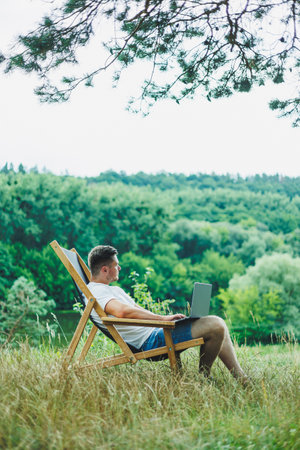 Young man lying on a chair in nature and working with a laptop, resting alone, thoughtfully looking up. A man on a trip, in the countryside. the concept of people's lifestyleの写真素材