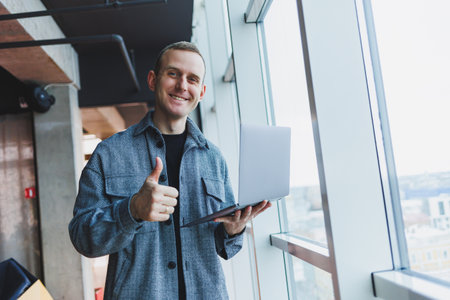 Portrait of a smart intelligent male manager, he is holding a laptop for research in the office, a business man in elegant clothes playing on the touchpad. Standing in the office by a large windowの写真素材