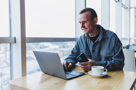 Successful happy business man is sitting at a table in a cafe, holding a cup of coffee and using a laptop.の写真素材