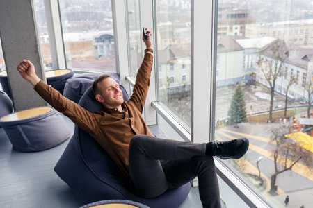 Young successful male businessman is sitting near a large window in the office and relaxing. Rest...