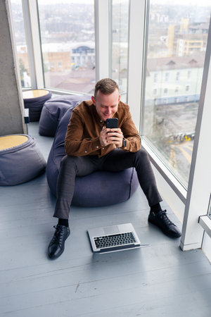 Young successful male businessman is sitting near a large window in the office and relaxing. Rest...