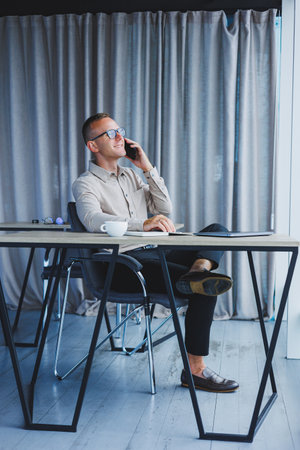 Indoor shot of cheerful bearded male freelancer work at distant, manages finances via smart phone,...