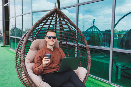 A businessman in formal wear sits on an armchair and looks at a laptop connected to a 4g wireless network on the terrace of a modern office while working remotelyの写真素材