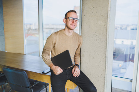 A businessman in glasses and stylish clothes drinks coffee in a modern workspace. A male freelancer...