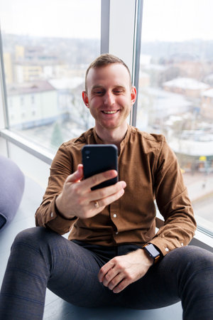 Handsome young male manager is sitting on the floor with the phone near the panoramic window. Male...