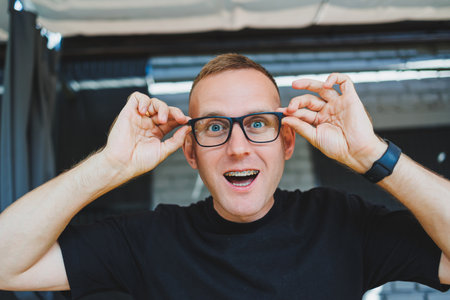 A cute young man in glasses and a black t-shirt stands on a summer terrace in a cafe and smiles...
