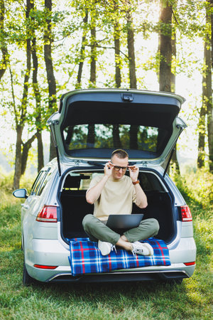 Successful man working outdoors from his car, sitting in the trunk and looking at the laptop. Online...